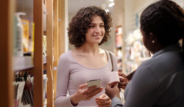 Young pretty customer with mobile phone in hand looking at female shop assistant and listening to description of new cosmetic product